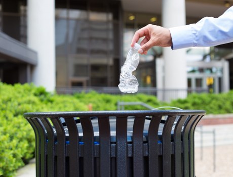 A person wearing a light blue long-sleeved shirt is seen disposing of a crumpled clear plastic water bottle into a black, cylindrical outdoor rubbish bin with vertical slats. The scene is outdoors, possibly in a courtyard or near a commercial building, with a background featuring blurred architectural elements and greenery. The ground surface appears to be paved, and the trash bin is positioned on a flat surface surrounded by garden plants and a concrete walkway. The image emphasizes waste disposal, aligning with rubbish removal services, and was taken in a setting that could be located in Watford or nearby areas of Hertfordshire. The focus is on the act of rubbish disposal, highlighting environmental responsibility in an urban environment, which is relevant to services provided by House Clearance Watford.
