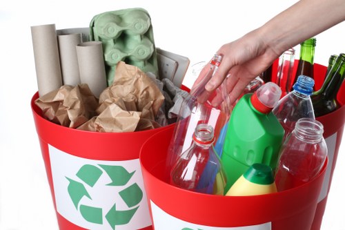 A close-up of two red recycling bins with white labels featuring green recycling symbols, filled with various household waste items. The left bin contains cardboard tubes, crumpled brown paper, and an egg carton, all made of paper or cardboard with textured surfaces. The right bin holds clear plastic bottles, some with caps, of different shapes and sizes, with smooth, glossy surfaces, alongside a green plastic bottle with a red cap and some glass bottles with green, brown, and clear hues. A person's hand is reaching into the right bin, indicating active waste sorting or collection. The background is plain and neutral, emphasizing the waste items and bins, which are positioned on a white surface. This image is representative of rubbish collection and recycling efforts typical to residential areas around Watford, reflecting environmentally conscious waste management practices as offered by local rubbish removal services like House Clearance Watford.