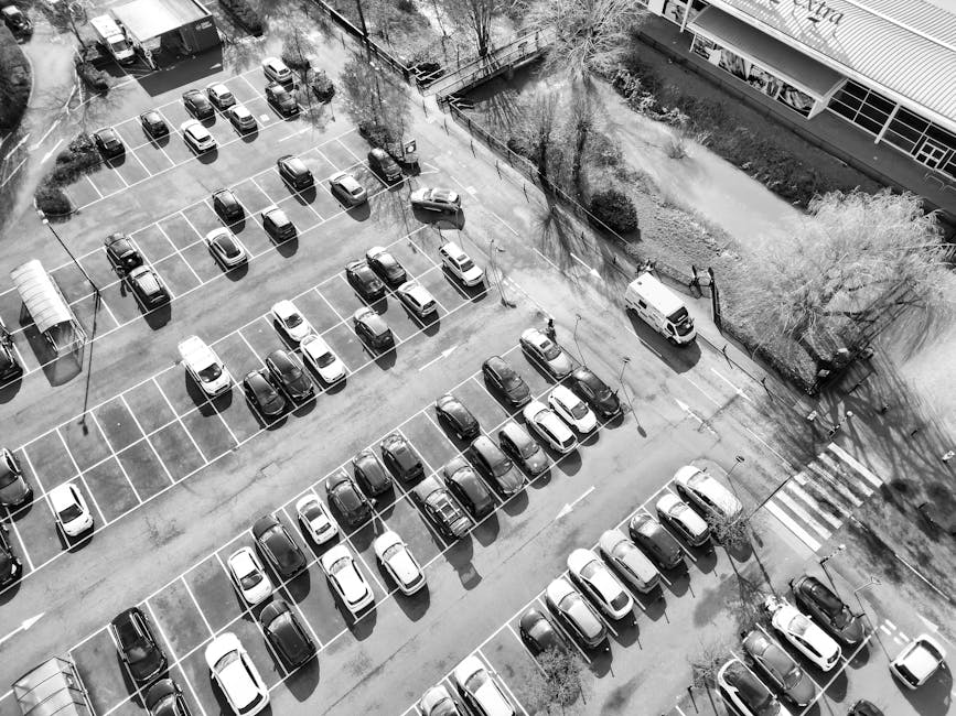 Aerial view of a large outdoor parking lot filled with numerous parked cars arranged in neat rows, predominantly in shades of white, black, and grey. The parking spaces are clearly marked with white l
