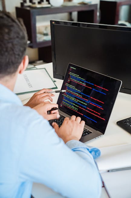 A person seated at a desk using a laptop with a black screen displaying multi-colored lines of computer code. The individual, seen from behind, has short dark hair and is wearing a light blue shirt. T