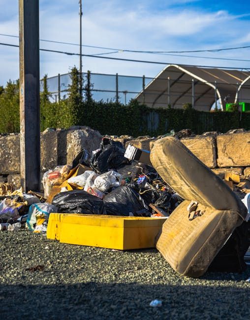 A large pile of black rubbish bags, made of plastic with a glossy finish, is stacked against a modern building with a tiled exterior wall composed of large beige tiles. The bags are bulging with waste